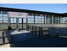 Eingang zum S-Bahnhof Bornholmer Straße mit einer Glasfassade und offenen Türen. Der Bereich ist von Metallgeländern umgeben, und der Himmel ist klar und blau.