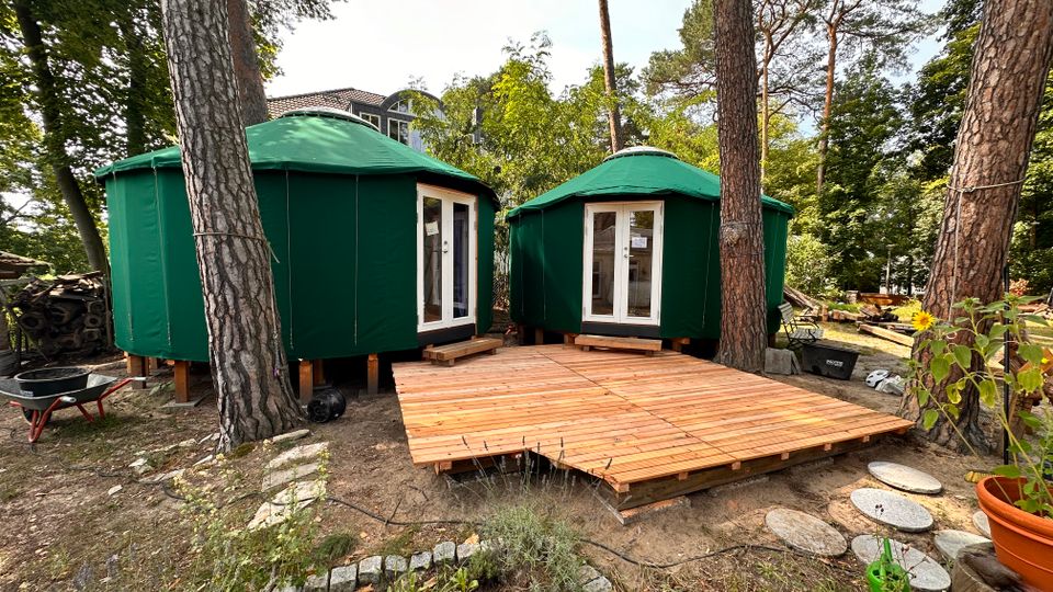 Zwei grüne, runde Zelte mit spitz zulaufenden Dächern stehen nebeneinander auf einem Waldgrundstück, umgeben von hohen Bäumen. Vor den Zelten liegt eine Holzterrasse, und im Vordergrund sind Gartenpflanzen sowie ein gepflasterter Weg sichtbar.