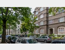 Straßenansicht mit mehrstöckigen Wohngebäuden aus braunem Putz und weißen Fenstern, ergänzt durch kleine Grünflächen und geparkte Autos entlang des Gehwegs. Im Vordergrund stehen Bäume, die teilweise die Gebäudeansicht verdecken.