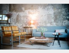 Wohnzimmer im industriellen Stil mit einer grauen Couch, zwei gelben Ledersesseln mit Metallrahmen und einem rustikalen Holztisch auf einem Teppich. Im Hintergrund eine Wand mit abgenutztem Putz, ein kleiner runder Beistelltisch mit Lampe und ein rechteckiger Wandspiegel.