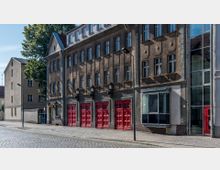 Straßenansicht eines historischen Gebäudes mit vier roten Garagentoren im unteren Bereich, vermutlich einer ehemaligen Feuerwache. Die Fassade ist schlicht gestaltet, mit hohen Fenstern und dezenten Verzierungen, und das Gebäude steht an einer gepflasterten Straße.