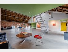 Großzügiger Loftraum in einem umgebauten Industriegebäude mit sichtbarer Holzbalkendecke, Backsteinwänden und Betonboden. Eine moderne Metalltreppe führt zu einer Galerie; im offenen Wohnbereich stehen Sitzmöbel und Tische, an den Wänden hängen Kunstwerke und es gibt mehrere niedrige Rundbogenfenster.