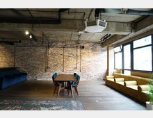 Industrie-Stil-Raum mit Backsteinwand, Holzboden und sichtbaren Rohrleitungen an der Decke. Ein Holztisch mit auffälligen Stühlen steht zentral, daneben eine lange gelbe Polsterbank vor großen Fenstern.