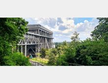 Ein großes, industrielles Stahlbauwerk steht zwischen dicht bewachsenen Bäumen, mit einem Weg im Vordergrund, auf dem Menschen spazieren. Im Hintergrund sind ein Fluss und eine weite Landschaft unter einem bewölkten Himmel sichtbar.
