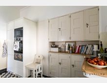 Helle Küche mit weißer Einbauküche, eingebautem Backofen und Mikrowelle auf der linken Seite. Rechts befinden sich beige Schränke und ein Holzarbeitsbereich, der mit Büchern, Geschirr und Obst dekoriert ist; der Boden ist in schwarz-weißen Karomustern gefliest.