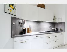 Moderne Küche mit weißen Fronten, grauer Arbeitsplatte und passendem Rückwandpanel in Betonoptik. Oberschränke mit integrierter Beleuchtung, Edelstahl-Spüle, Kochfeld, Wandhalterung mit Küchenutensilien und dekorative Elemente wie Pflanzen und eine Zitronen-Illustration ergänzen den Raum.