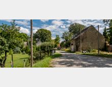 Ländliche Straßenszene mit gepflastertem Weg, umgeben von Gärten mit Bäumen und Sträuchern. Auf der rechten Seite ein altes, mit Efeu bewachsenes Gebäude aus Backstein und links ein Drahtzaun, dahinter eine gepflegte Wiese mit weiteren Pflanzen.