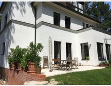 Einfamilienhaus mit weißer Fassade, großen dunklen Fenstern und einem flachen Anbau im Garten. Vor der Terrasse stehen Topfpflanzen und Gartenmöbel, daneben ein geschlossener Sonnenschirm.