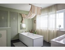 Modernes Badezimmer mit einer freistehenden weißen Badewanne, hellgrünen Wänden und hängenden Stoffvorhängen als dekoratives Element. Großes Fenster mit halbtransparenten Vorhängen, schwarzer Boden und schlichtes Wanddesign.