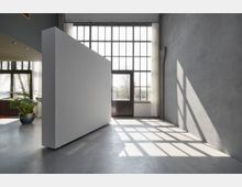 Minimalistisch gestalteter Innenraum mit hohen Decken und großen, bodentiefen Fenstern, die viel Tageslicht hereinlassen. Der Raum hat eine graue Betonwand, einen weißen freistehenden Block und einen Pflanzentopf in der linken Ecke.