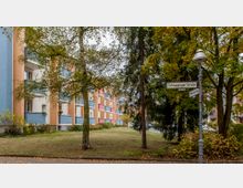 Wohngebäude mit bunten Balkonen in einer Wohnstraße, umgeben von Bäumen und einer kleinen Grünfläche. Im Vordergrund stehen ein Straßenschild für die „Schöppinger Straße“ und eine nostalgisch gestaltete Laterne.