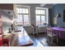 Helle Küche mit Holzboden und zwei großen Fenstern, die viel Tageslicht hereinlassen. Auf der linken Seite befindet sich eine lange Arbeitsplatte mit Spüle, rechts ein Esstisch mit violettem Tischtuch und Stühlen, dazu eine blau gestrichene Wand und ein Heizkörper unter den Fenstern.