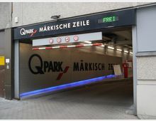 Einfahrt zu einem Parkhaus mit der Aufschrift „Q-Park Märkische Zeile“, ausgestattet mit einer Schranke, LED-Beleuchtung und Informationen zu Höhen- und Geschwindigkeitsbegrenzungen. Rechts ist ein Ticketautomat zu sehen, und oberhalb des Eingangs wird mit einem grünen Schild angezeigt, dass freie Plätze verfügbar sind.