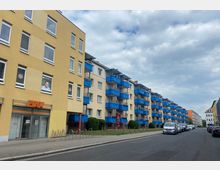 Straßenansicht mit Wohngebäuden aus mehreren Stockwerken, deren gelbe Fassaden durch blaue Balkone akzentuiert sind. Im Vordergrund ist ein Geschäft mit der Beschriftung „DAK“ zu sehen, während Autos entlang der gepflasterten Straße geparkt sind.