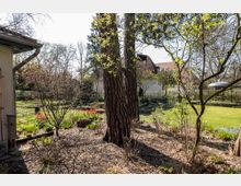 Ein Garten mit Bäumen und Sträuchern im Vordergrund, darunter vereinzelt rote Tulpen. Im Hintergrund befinden sich ein Rasenbereich, eine weiß gestrichene Villa mit rotem Ziegeldach sowie ein kleiner Pavillon.
