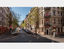 Straßenszene mit gepflasterter Nebenstraße, umgeben von Altbauten mit reich verzierten Fassaden. Im Erdgeschoss des Eckgebäudes befindet sich das "Café Gediz", daneben geparkte Autos und Fahrräder; Bäume säumen die Straße, teils mit frischem Laub.