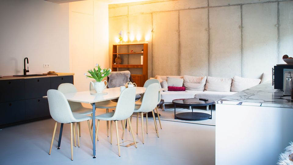 Offener Wohn- und Küchenbereich im modernen Industriestil mit Betonwand, schwarzer Küchenzeile und heller Kücheninsel. Davor stehen ein Esstisch mit vier Stühlen sowie eine große helle Eckcouch mit rundem Couchtisch und Regal im Hintergrund.