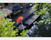 Holzterrasse mit Sitzgelegenheiten und einem gedeckten Tisch, direkt an einem kleinen Kanal gelegen, an dem ein langes, schmales Boot vertäut ist; im Vordergrund befinden sich blühende rote Blumen und grüne Blätter.