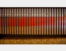 Ansicht eines modernen Innenraums mit roter Wand und gefliesten Böden, durch eine schwarze Metallgitterstruktur im Vordergrund betrachtet. Die Beleuchtung im oberen Bereich erzeugt eine gleichmäßige, warme Ausleuchtung.