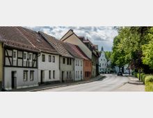 Straßenszene in einem kleinen Dorf oder einer Kleinstadt mit historischen Häusern im Fachwerkstil, teils mit verputzten Fassaden, entlang einer kurvigen Straße. Rechts säumen Bäume mit dichtem Grün den Gehweg, während im Hintergrund weitere weiße Gebäude sichtbar sind.