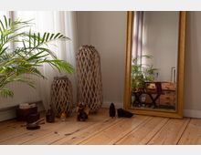 Helle Zimmerecke mit breitem Holzdielenboden, bodentiefem Fenster mit transparenten Vorhängen und einer großen Zimmerpflanze. Daneben stehen zwei geflochtene, hohe Lampenschirme oder Körbe und ein großer, goldgerahmter Standspiegel, der eine Kommode und weitere Pflanzen im Raum reflektiert.
