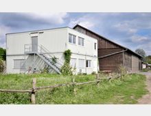 Zweistöckiges Containergebäude mit Metalltreppe und rankenden Pflanzen, daneben eine Scheune aus Holz. Im Vordergrund eine Wiese mit Wildblumen und einem Holzzaun, im Hintergrund teilweise bewölkter Himmel.