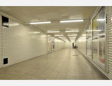 Ein heller unterirdischer Fußgängertunnel mit weißen Fliesenwänden und einer beleuchteten Decke. Entlang einer Wand befinden sich Schaukästen mit Plakaten, und ein Mann steht in der Ferne neben einem Reinigungswagen.