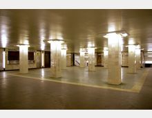 Unterirdische U-Bahn-Passage mit weiß gefliesten Säulen, hellem künstlichem Licht und einer offenen Bodenfläche; im Hintergrund befinden sich Hinweise auf Geschäfte sowie Treppen und Rolltreppen.