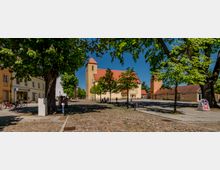Gepflasterter Dorfplatz mit großen schattenspendenden Bäumen und Sitzbänken, umgeben von niedrigen Gebäuden. Im Hintergrund steht eine helle Kirche mit rotem Satteldach und Turm unter blauem Himmel.