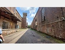 Blick auf eine schmale, gepflasterte Gasse zwischen alten Backsteinfabrikhallen mit großen Fenstern und verwitterten Wänden, die an ein Industriegebiet erinnern. Im Hintergrund erhebt sich ein höheres Gebäude, und am Ende der Gasse sind Bäume sichtbar unter einem teilweise bewölkten Himmel.