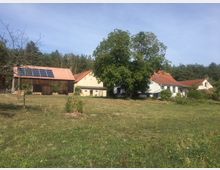 Ländliche Szene mit mehreren Gebäuden, darunter eine Scheune mit Solarpaneelen auf dem Dach, umgeben von Wiesen und Bäumen. Im Hintergrund ist ein Wald sichtbar, der den Ort umrahmt.