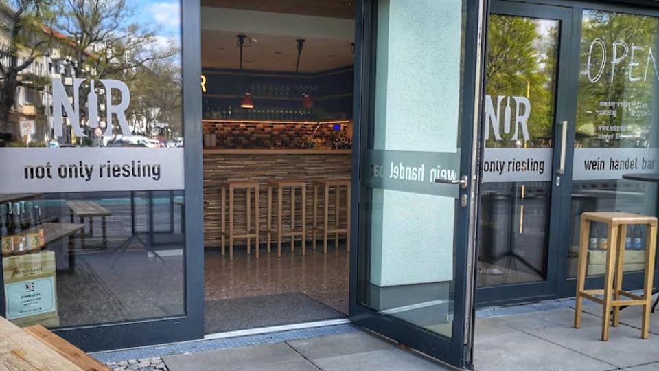 Eingangsbereich einer modernen Weinbar mit großen Glasfronten und geöffneten Glastüren, beschriftet mit „NOR – not only riesling“ und „OPEN“. Durch die Türen sind eine lange Bar, hohe Holzhocker und Regale mit Flaschen im Innenraum sichtbar.