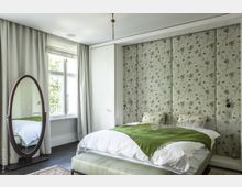 Schlafzimmer mit einem Doppelbett, das mit einem grünen Überwurf und floral gemusterten Kissen bedeckt ist. Die Wand hinter dem Bett ist mit gepolsterten Paneelen mit Blumenmuster gestaltet, ein großes ovales Spiegel steht links neben einem Fenster mit weißen Vorhängen.