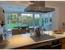 Moderne Wohnküche mit Kochinsel im Vordergrund, auf der ein Kochfeld, Gewürze und eine Obstschale platziert sind. Im Hintergrund ein Essbereich mit großen Fenstern und Blick in einen gepflegten Garten mit Rasen und Bäumen.