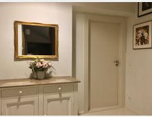 Innenraum mit einer weißen Kommode mit Holzplatte, auf der ein Blumenarrangement steht. Darüber hängt ein rechteckiger Spiegel mit goldener Rahmung, daneben eine geschlossene Zimmertür und ein gerahmtes Wandbild.