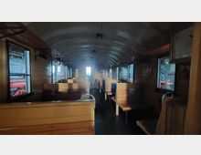 Innenraum eines historischen Zugwaggons mit Holzausstattung, Sitzbänken auf beiden Seiten und mehreren Fenstern, durch die Außenansichten sichtbar sind. Der Waggon hat eine gewölbte Decke und eine Tür am Ende des Ganges.