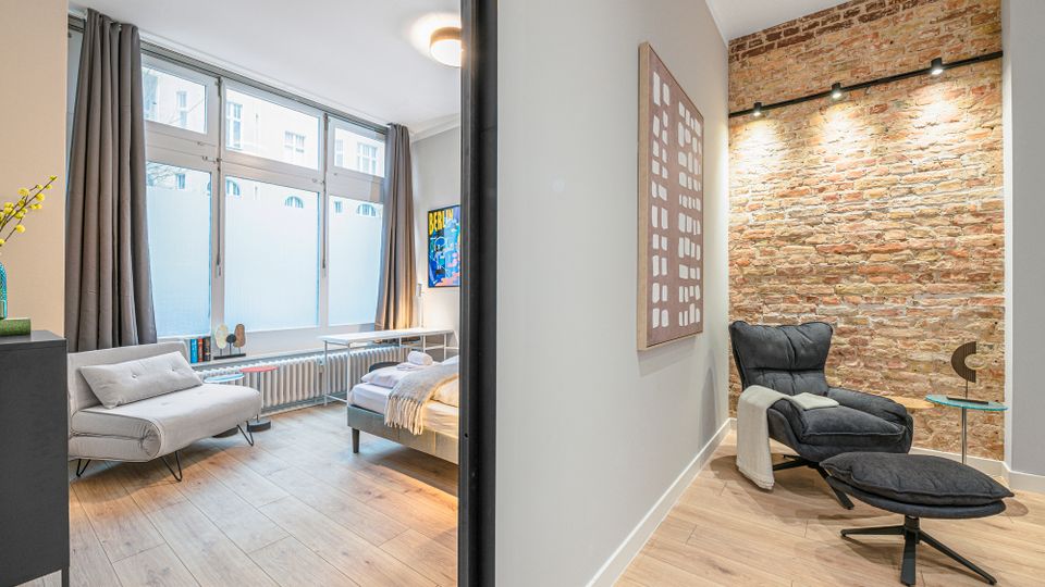 Modernes Apartmentzimmer mit großen Fenstern und grauen Vorhängen, hellem Holzboden sowie Bett und kleinem Sofa. Rechts daneben eine Sitzecke mit schwarzem Lounge-Sessel, Beistelltisch und freiliegender Ziegelwand mit Schienenbeleuchtung.