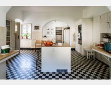 Helle Küche mit einem schwarz-weiß karierten Boden, einer zentralen Kochinsel mit Holzarbeitsplatte, und weißen Wänden. An den Seiten befinden sich Einbauschränke, offene Regale und Möbel; ein Bogen verbindet zwei Bereiche. Fensterscheiben mit Streben sorgen für Tageslicht.