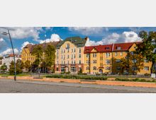 Straßenansicht mit mehreren mehrfarbigen Wohngebäuden im Stil früherer Baujahre, hauptsächlich in Gelb- und Rottönen mit weißen Fensterrahmen. Im Vordergrund befindet sich ein Bürgersteig, eine kleine Grünfläche mit Blumenbeeten sowie ein gepflasterter Straßenbereich; einzelne Bäume sind entlang der Gebäude gepflanzt.