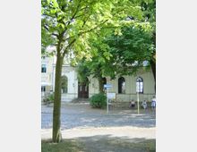 Historisches Gebäude mit heller Fassade und großen Rundbogenfenstern, teils verdeckt durch Bäume im Vordergrund; davor ein gepflasterter Platz mit einigen Menschen, die auf einer Bank sitzen.