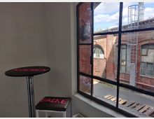 Ein Innenraum mit einem hohen Tisch und einem Hocker, beide mit pinkfarbenen Schriftzügen ("Kuchen" und "Backe") versehen. Durch das große, industrielle Fenster ist eine Backsteinfassade mit Metalltreppe und alten Fenstern zu sehen, sowie Paletten vor der Fensterbank.