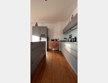 Moderne Küche mit grauen Hochglanz-Schränken und Arbeitsflächen auf beiden Seiten eines schmalen Gangs, einem Holzfußboden und einem kleinen Holzschrank im Hintergrund. Auf einem der hohen Schränke sitzt eine Katze.