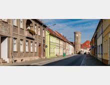 Straße in einem historischen Stadtzentrum mit Reihenhäusern auf beiden Seiten, teils renoviert, teils mit alten Fassaden. Im Hintergrund steht ein runder, steinerner Uhrturm mit rotem Dach.