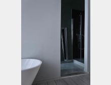 Modernes Badezimmer mit einer freistehenden weißen Badewanne auf hellem Holzboden. Im Hintergrund befindet sich eine offene Tür mit Blick auf eine Dusche mit Glaswänden und einen schmalen Spiegel, der an die Wand gelehnt ist.