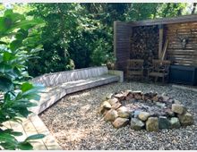 Gartenbereich mit Feuerstelle aus Steinen, umgeben von einer Kiesfläche. Eine gebogene Holzbank, zwei Holzstühle und eine Wand aus gestapeltem Holz sowie eine Holztrennwand mit Geweihdeko prägen die Szenerie, umrahmt von dichtem Grün.
