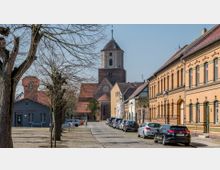 Straßenszene in einer historischen Altstadt mit gepflasterter Straße, geparkten Autos und Bäumen. Im Hintergrund eine Kirche aus Backstein mit hohem Turm und umliegenden Gebäuden mit Ziegeldächern.