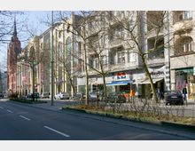 Straßenszene mit mehrstöckigen Gebäuden, die Geschäfte und Cafés im Erdgeschoss haben, sowie einer Straße mit parkenden und fahrenden Autos. Im Hintergrund erhebt sich ein großes rotes Gebäude mit einem Kirchturm, Bäume ohne Laub säumen das Bild.