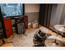 Probeaufnahmeraum mit einem Schlagzeug im Vordergrund, Verstärkern und Mikrofonen auf einem Teppichboden. Im Hintergrund ist durch ein Fenster ein separater Bereich mit weiteren Instrumenten erkennbar.
