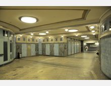 Unterirdischer Fußgängertunnel mit hellen Fliesenwänden in verschiedenen Blautönen, eingerahmt von dekorativen Elementen. Die Decke ist mit runden und rechteckigen Leuchten ausgestattet, und der Boden ist mit gemusterten Steinfliesen ausgelegt; im Hintergrund ein Ausgangsbereich mit Wegweiser.