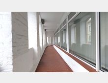 Überdachter Korridor mit weißen Ziegelwänden und großen Glasfenstern auf der rechten Seite, die von vertikalen Jalousien verdeckt sind; der Boden ist mit rotbraunen Fliesen ausgelegt.